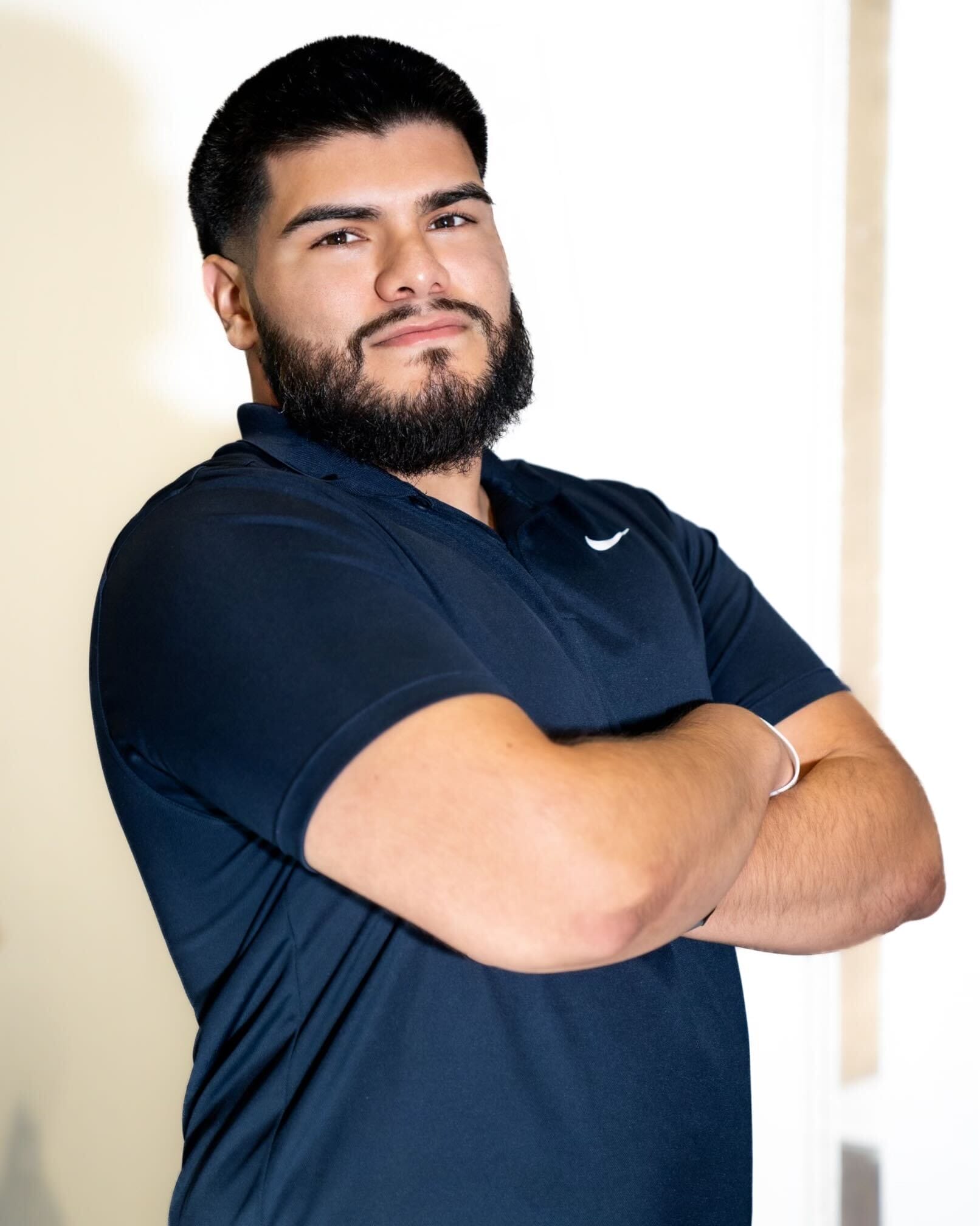 A man with a beard and mustache wearing a black polo shirt stands with his arms crossed.