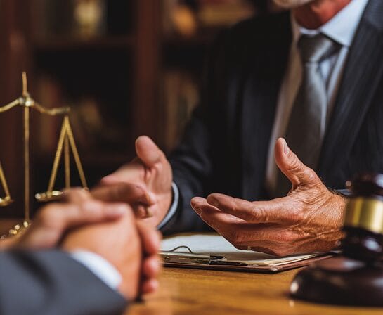 Two people in business attire sit at a desk discussing legal matters; a gavel, legal scales, and documents are visible in the foreground.